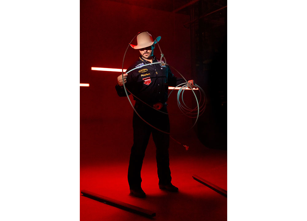 Anthony Lucia vistiendo una camisa negra y un sombrero de vaquero, sosteniendo una cuerda con iluminación roja detrás de él.